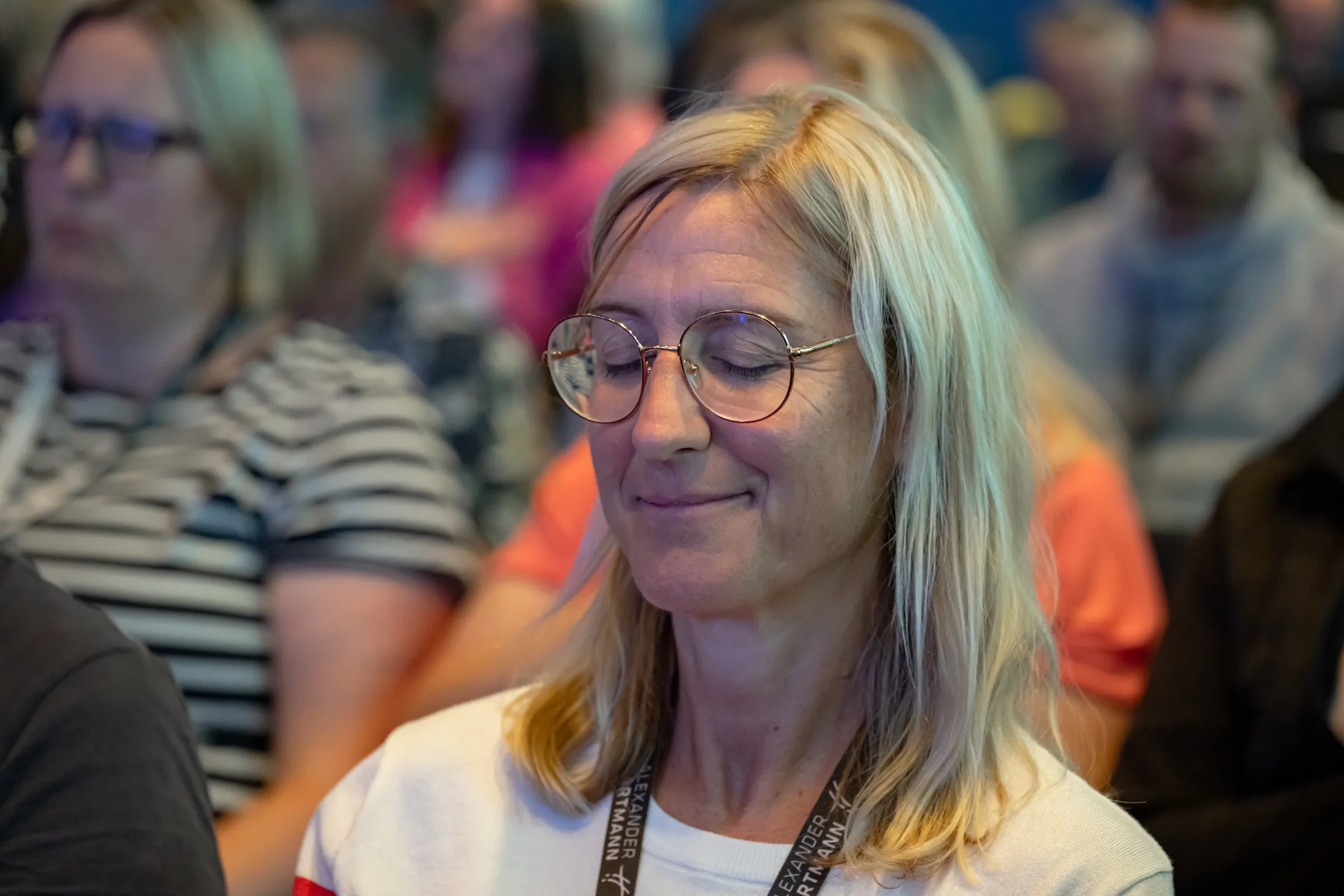 Seminarteilnehmerin mit Brille sitzt mit geschlossenen Augen und leichtem Lächeln in einer Hypnoseübung, hinter ihr der volle Saal